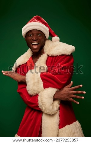 overjoyed african american man in santa claus costume gesturing and looking at camera on green