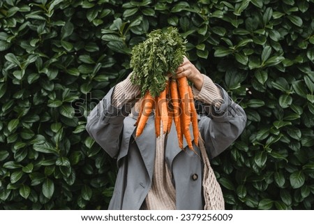 Smiling woman with fresh organic carrots and mesh bag against green leaf wall. Concept of people doing sustainable shopping of eco local products without plastic packaging. Minimalist lifestyle