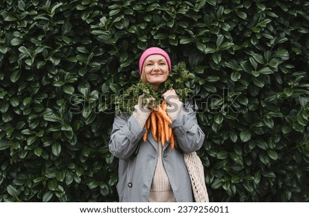 Smiling woman with fresh organic carrots and mesh bag against green leaf wall. Concept of people doing sustainable shopping of eco local products without plastic packaging. Minimalist lifestyle