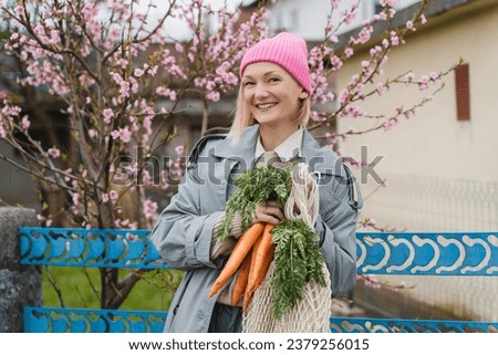 Young smiling woman with bouquet of fresh organic carrots and mesh bag in garden outside. People doing sustainable shopping of regional products from local market without plastic packaging