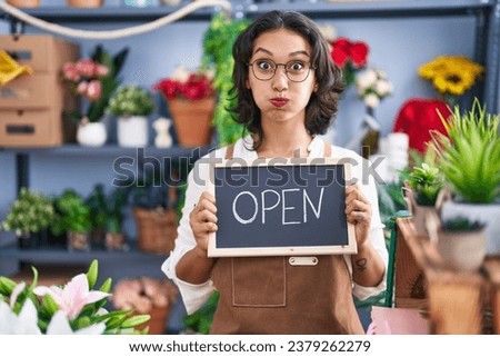 Young hispanic woman working at florist holding open sign puffing cheeks with funny face. mouth inflated with air, catching air. 
