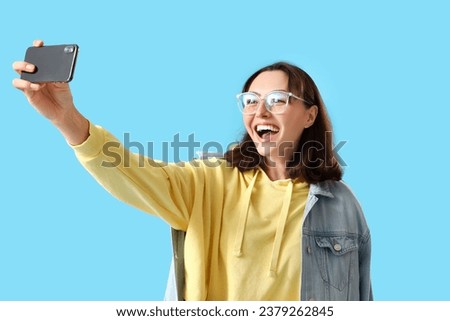 Young woman in yellow hoodie with mobile phone taking selfie on blue background