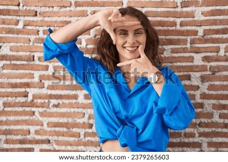 Beautiful brunette woman standing over bricks wall smiling making frame with hands and fingers with happy face. creativity and photography concept. 