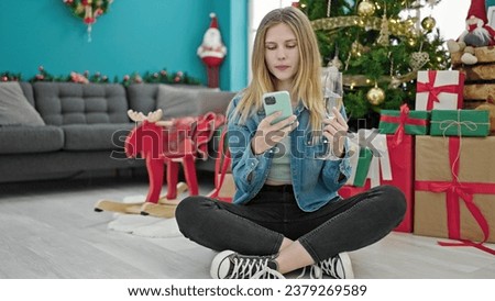 Young blonde woman celebrating christmas holding champagne using smartphone at home