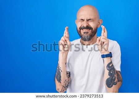 Hispanic man with tattoos standing over blue background gesturing finger crossed smiling with hope and eyes closed. luck and superstitious concept. 