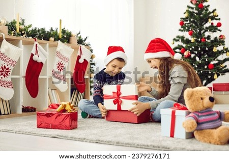 Overjoyed small children in santa hats have fun unpack wrapped presents on merry Christmas morning near fir tree. Smiling kids open gifts from Santa Claus on New Year eve on winter holiday.