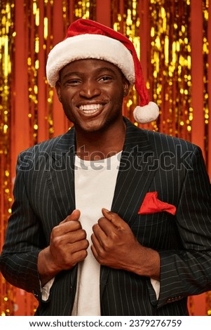 happy african american man in santa hat and black blazer smiling on shiny background with tinsel