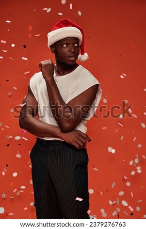 young african american man in white tank top and santa hat looking away under confetti on red