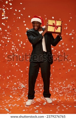 cheerful african american man in elegant suit and santa hat holding present under confetti on red