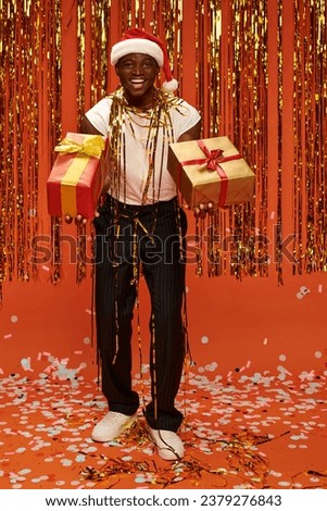 african american man in santa cap holding gifts and looking at camera near christmas decor on red