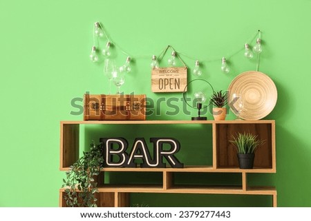 Wooden shelving unit with word BAR, decorative sign WELCOME WE'RE OPEN and lamps near green wall