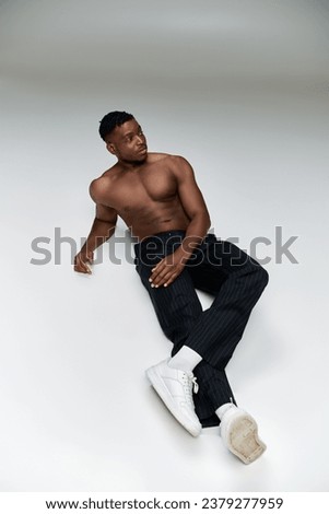 confident shirtless african american fitness model in black pants sitting and looking away on grey