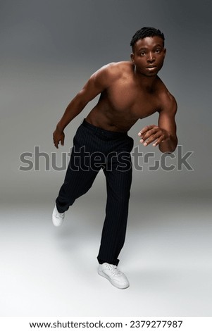 shirtless and sportive african american man in black pants posing on one leg on grey backdrop