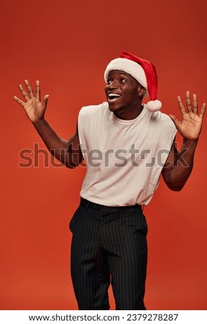 amazed and overjoyed african american man in santa hat showing wow gesture and looking away on red