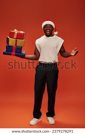 excited african american guy in santa cap holding gift boxes and showing wow gesture on red backdrop