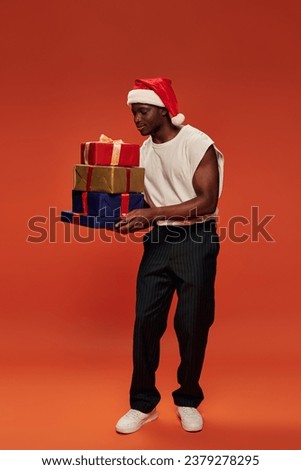 pleased african american man in festive santa hat holding colorful christmas presents on red