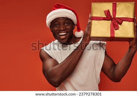 overjoyed african american guy in santa hat holding gift box and looking at camera on red backdrop