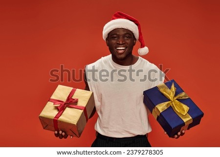 joyful african american man in christmas hat and white tank top posing with festive presents on red