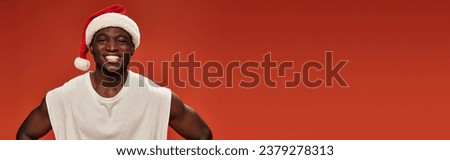 joyful african american guy in santa hat and white tank top looking at camera on red, banner