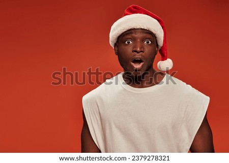 shocked african american guy in santa hat looking at camera with open mouth on red background