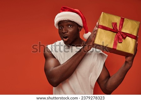 surprised african american man in santa hat holding gift box and looking at camera on red backdrop