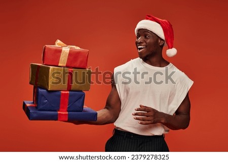 amazed and overjoyed african american man in santa hat looking at colorful gift boxes on red