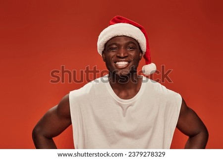 cheerful african american man with radiant smile posing in santa hat and looking at camera on red