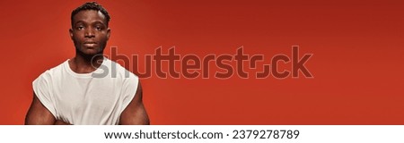 confident dark skinned man in white tank top looking at camera on red studio backdrop, banner