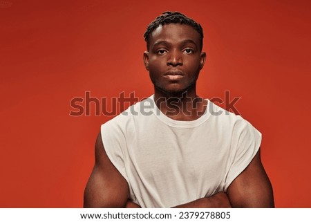 confident athletic african american man in white tank top looking at camera on red studio backdrop