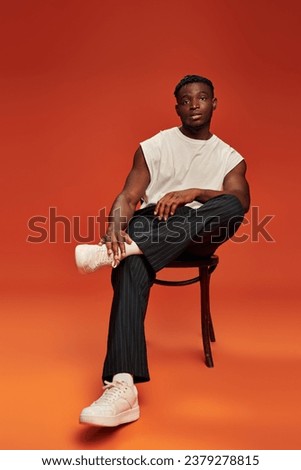 african american male model in stylish street wear sitting on chair on red and orange backdrop