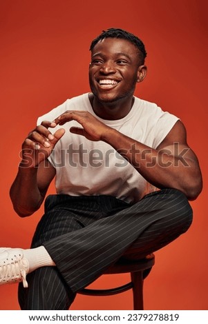 laughing african american man in white tank top sitting on chair and gesturing on red backdrop