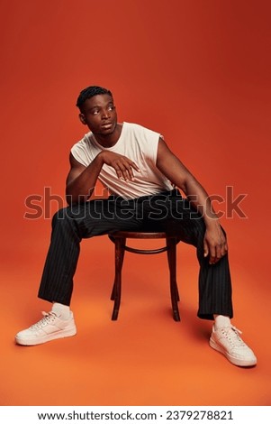 modern and stylish african american man sitting on chair and looking away on red and orange backdrop