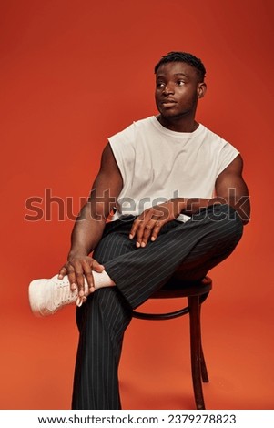 hot african american guy in black pants and white tank top posing on chair and looking away on red