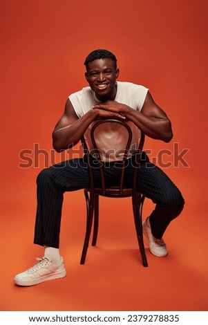 excited african american man in white tank top and pants sitting on chair on red and orange backdrop