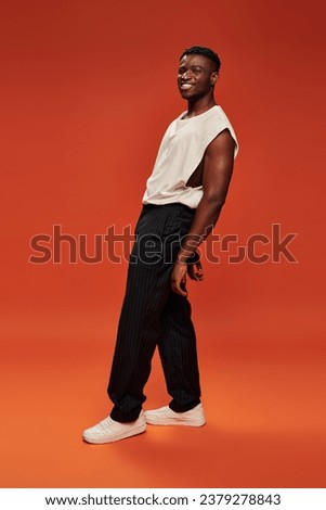 carefree african american man in black pants and white tank top posing on red and orange backdrop