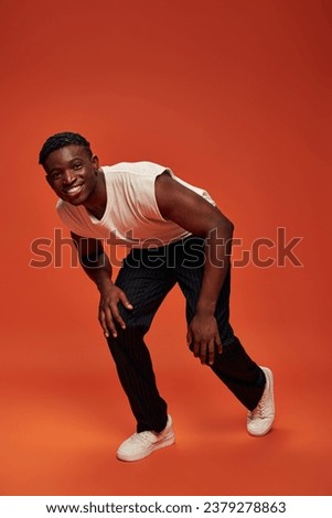 happy african american guy in white tank top and pants leaning forward and looking at camera on red
