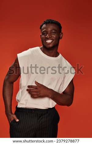 joyful african american man with hand in pocket of black pants looking at camera on red backdrop