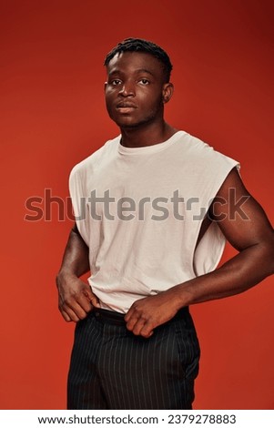 pensive african american man in white tank top and black pants looking away on red backdrop