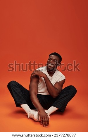 excited stylish african american man sitting and laughing with closed eyes, red and orange backdrop