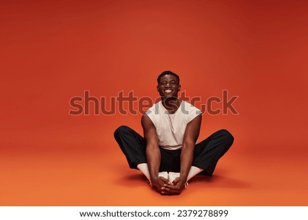 overjoyed african American man in white tank top and black pants sitting on red and orange backdrop