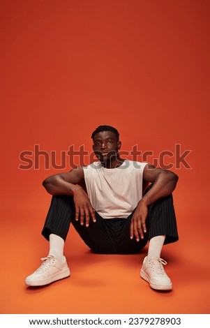 confident and sportive african american man sitting and looking at camera on red and orange backdrop