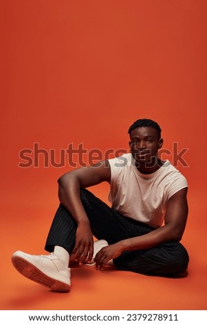 african american man in trendy casual wear sitting and looking at camera on red and orange backdrop