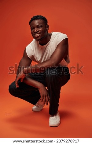 carefree african american man in trendy street wear sitting on haunches on red and orange backdrop
