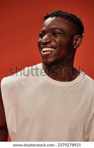 excited african american man in white tank top laughing and looking away on red backdrop, happiness