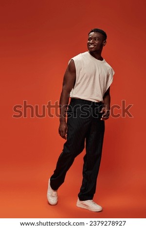 joyous and hot african american man in white tank top and black pants looking away on red backdrop