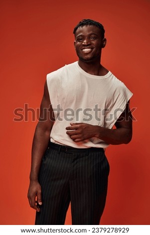 cheerful african american guy in white tank top and black pants looking at camera on red backdrop