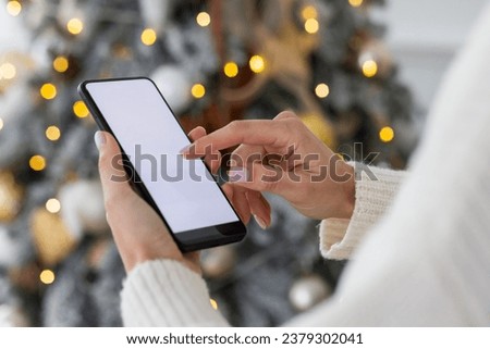 Close-up photo of young female hands holding and using a mobile phone, typing on the screen with a finger, white mock-up on a background of a decorative Christmas tree.