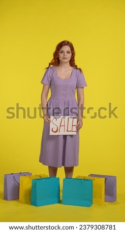 An attractive young woman is standing surrounded by shopping bags and holding a banner that says Sale. A redhaired woman stands full length in a studio against a yellow background. Vertical shot.