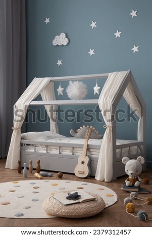 Aesthetic composition of cozy kid room interior with stylish bed, blue wall, braided pouf, round rug, books, colorful garland, gray curtain and personal accessories. Home decor. Template.
