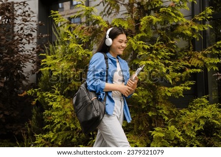 Beautiful young asian woman student in headphones smiling while walking in university campus